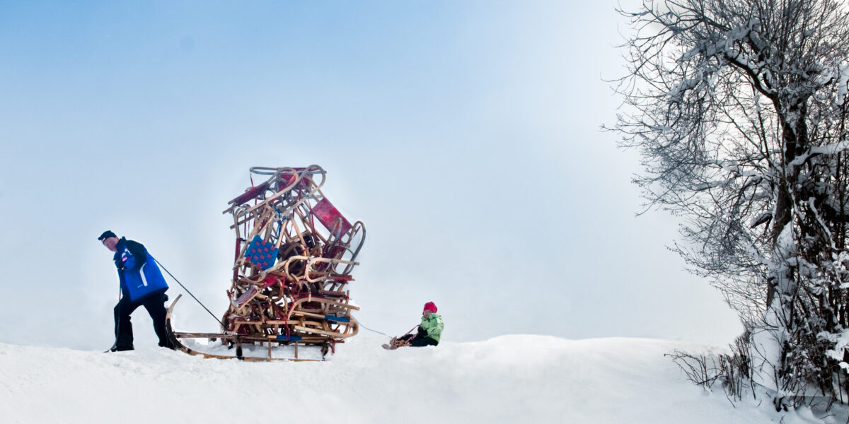 Eine Person zieht einen mit Geweihen beladenen Schlitten durch den Schnee, während ein Kind in Winterkleidung auf einem kleineren Schlitten folgt. Rechts steht ein schneebedeckter Baum.
