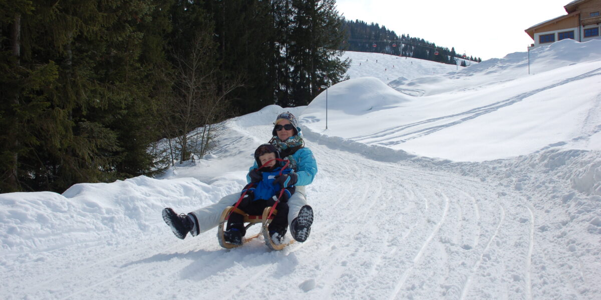 Ein Erwachsener und ein Kind fahren auf einem Schlitten einen verschneiten Hügel hinunter, umgeben von Kiefern und schneebedeckten Hängen unter einem teilweise bewölkten Himmel.
