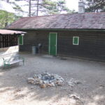 Eine Holzhütte mit einer grünen Tür und kleinen Fenstern, einer Feuerstelle aus Stein davor, einer Bank und einem Schuppen neben dem Gebäude in einem Waldgebiet.