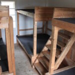 Ein kleines Zimmer mit vier hölzernen Etagenbetten, jedes mit einer schwarzen Matratze; durch ein Fenster im Hintergrund fällt natürliches Licht ein.