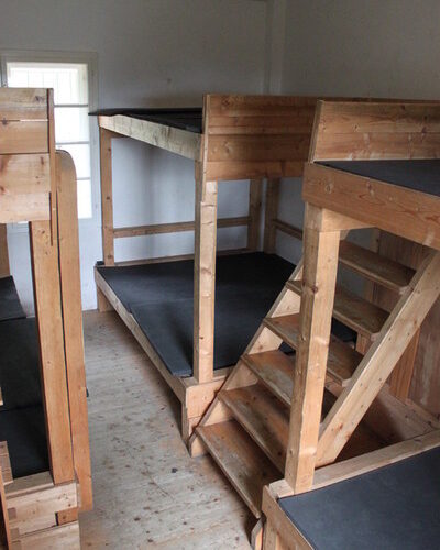 Ein kleines Zimmer mit vier hölzernen Etagenbetten, jedes mit einer schwarzen Matratze; durch ein Fenster im Hintergrund fällt natürliches Licht ein.