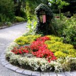 Geschwungener Kiesweg neben einem kreisförmigen Blumenbeet mit roten, gelben und weißen Blumen, mit einem Wappenschild inmitten von grünem Laub.