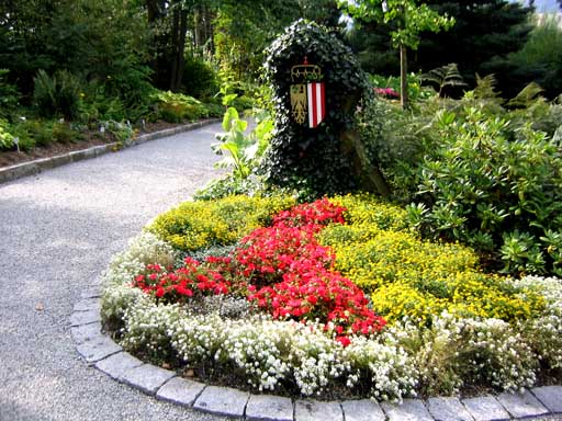 Geschwungener Kiesweg neben einem kreisförmigen Blumenbeet mit roten, gelben und weißen Blumen, mit einem Wappenschild inmitten von grünem Laub.