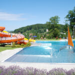 Freibad mit Wasserrutsche, kleinen Springbrunnen und einer segelförmigen Struktur, umgeben von Grün und Hügeln unter strahlend blauem Himmel.