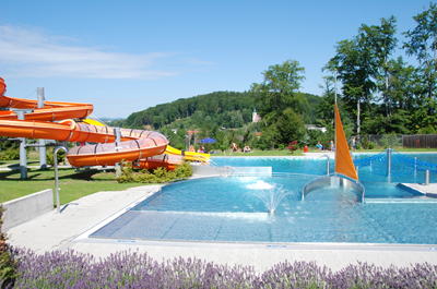 Freibad mit Wasserrutsche, kleinen Springbrunnen und einer segelförmigen Struktur, umgeben von Grün und Hügeln unter strahlend blauem Himmel.