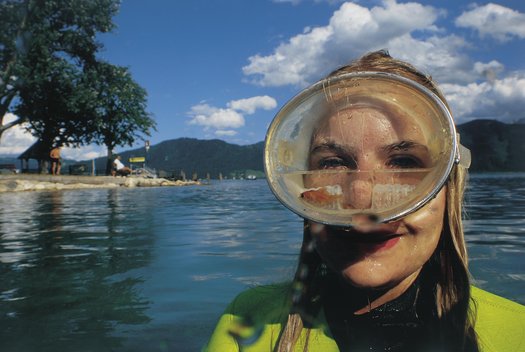 Eine Person in einem grünen Neoprenanzug steht lächelnd in einem See und trägt eine große Tauchermaske, die ihr Gesicht bedeckt; im Hintergrund sind Bäume, Berge und Menschen zu sehen.