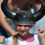 Junges Mädchen, das einen schwarzen Helm mit Hörnern im Wikinger-Stil trägt, lächelt und die Hände hebt, mit unscharfen Menschen im Hintergrund.