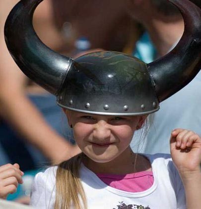Junges Mädchen, das einen schwarzen Helm mit Hörnern im Wikinger-Stil trägt, lächelt und die Hände hebt, mit unscharfen Menschen im Hintergrund.