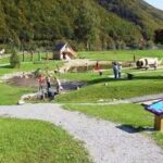 Ein kleiner Park mit Graswegen, Hinweisschildern, einem Teich, Holzkonstruktionen und mehreren Personen, die im Freien spazieren oder stehen.
