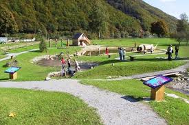 Ein kleiner Park mit Graswegen, Hinweisschildern, einem Teich, Holzkonstruktionen und mehreren Personen, die im Freien spazieren oder stehen.