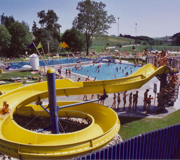 Öffentliches Freibad mit einer großen gelben Wasserrutsche, umgeben von Menschen, die sich auf einer Wiese sonnen, schwimmen und entspannen.