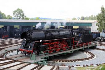 Auf einer runden Drehscheibe vor einem Ringlokschuppen steht eine schwarze Dampflokomotive, aus deren Schornstein Dampf aufsteigt.