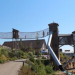 Die Kinder laufen über eine Seilbrücke und benutzen große Rutschen auf einem hölzernen Spielplatz unter einem strahlend blauen Himmel.
