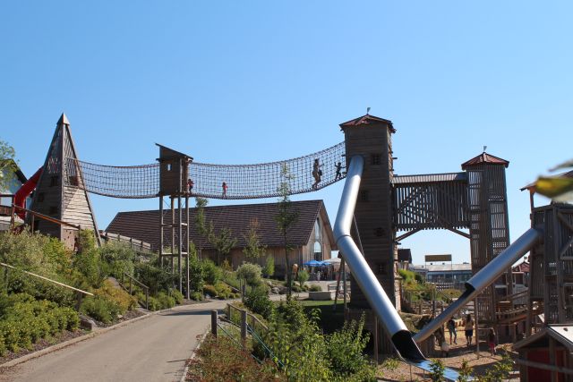 Die Kinder laufen über eine Seilbrücke und benutzen große Rutschen auf einem hölzernen Spielplatz unter einem strahlend blauen Himmel.