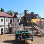 Kinderspielplatz im Freien mit einem Spielhaus, Holzkonstruktionen, einem grünen Spielwagen und Klettergeräten an einem sonnigen Tag.