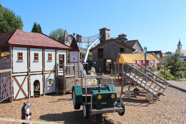 Kinderspielplatz im Freien mit einem Spielhaus, Holzkonstruktionen, einem grünen Spielwagen und Klettergeräten an einem sonnigen Tag.