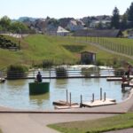 Ein kleiner Teich mit hölzernen Stegen und einer Person in einem runden Boot; ein Kind und ein Kinderwagen sind auf einem Weg zu sehen, mit Häusern und grasbewachsenen Hügeln im Hintergrund.