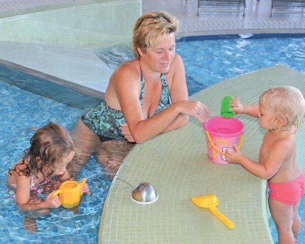 Ein Erwachsener und zwei kleine Kinder spielen mit Plastikspielzeug am Rande eines Schwimmbeckens. Der Erwachsene sitzt im Wasser, während die Kinder einen Eimer und Becher benutzen.