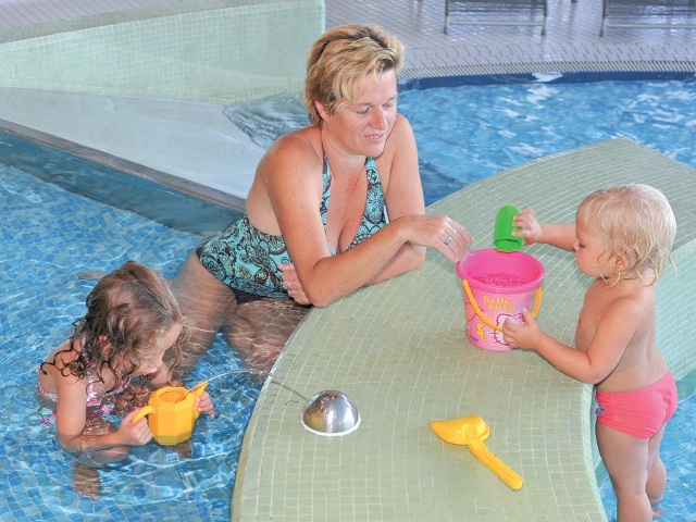 Ein Erwachsener und zwei kleine Kinder spielen mit Plastikspielzeug am Rande eines Schwimmbeckens. Der Erwachsene sitzt im Wasser, während die Kinder einen Eimer und Becher benutzen.