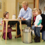 Zwei Erwachsene und zwei kleine Kinder sitzen um einen kleinen Tisch mit Molkereigeräten, darunter eine Milchkanne, in einem Raum mit Lehrtafeln an der Wand.