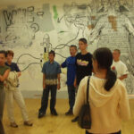 Eine Gruppe junger Leute steht in einem Raum einer Kunstgalerie mit Graffiti und großen Wandzeichnungen an den Wänden.