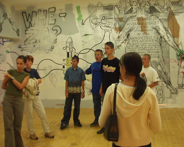 Eine Gruppe junger Leute steht in einem Raum einer Kunstgalerie mit Graffiti und großen Wandzeichnungen an den Wänden.