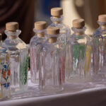 Eine Reihe von Klarglasflaschen mit Holzkorken, einige mit bunten Blumenmustern verziert, auf einer weißen Fläche ausgestellt.