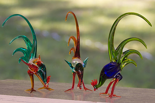 Drei bunte Glashahnfiguren mit langen Schwänzen werden auf einer flachen Oberfläche mit verschwommenem grünem Hintergrund ausgestellt.