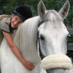 Ein Kind, das einen Helm trägt, umarmt den Hals eines weißen Pferdes und sitzt im Sattel. Im Hintergrund sind Bäume und ein Holzzaun zu sehen.