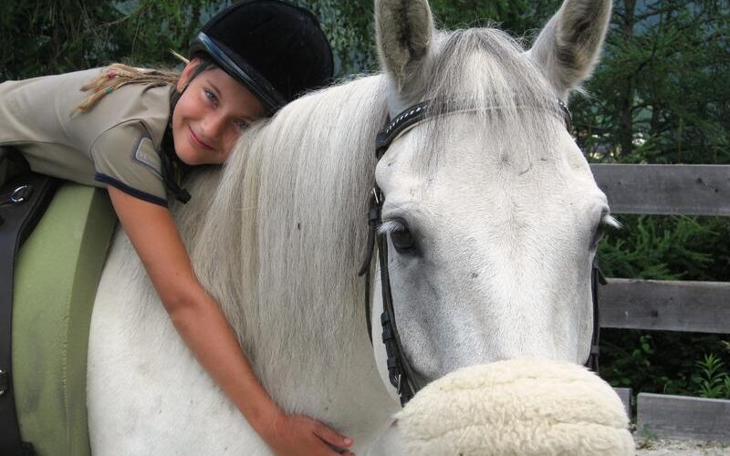 Ein Kind, das einen Helm trägt, umarmt den Hals eines weißen Pferdes und sitzt im Sattel. Im Hintergrund sind Bäume und ein Holzzaun zu sehen.
