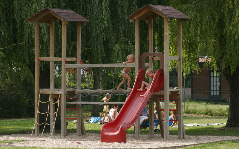 Kinder spielen auf einem hölzernen Spielplatz mit einer roten Rutsche, einer Strickleiter und einer Verbindungsbrücke in einem grasbewachsenen Parkgelände.