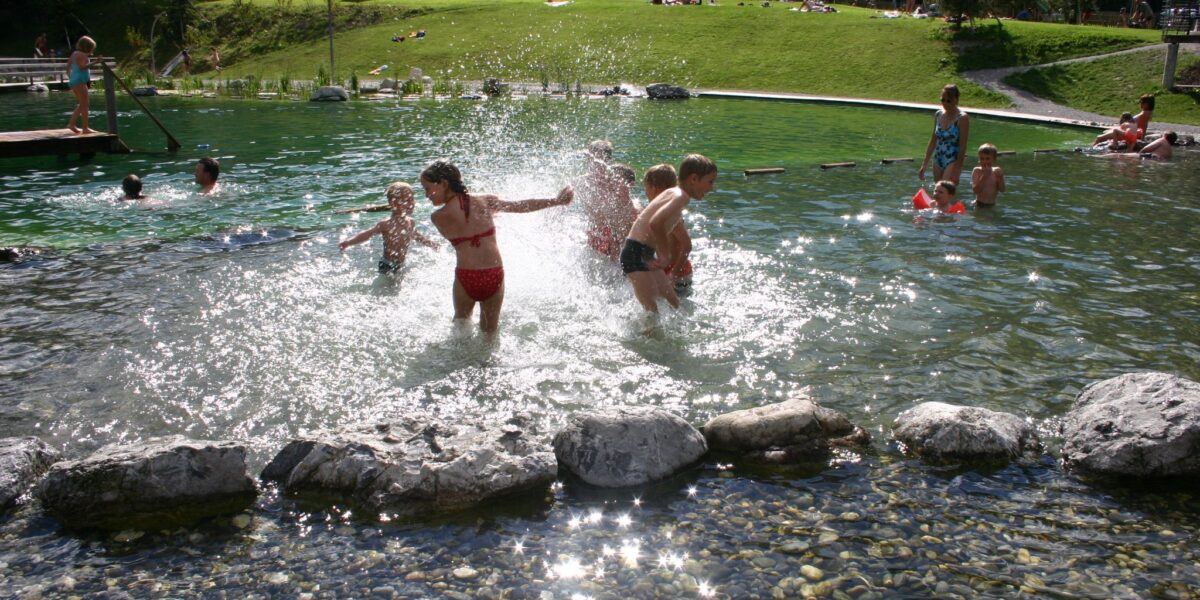 Kinder spielen und planschen in einem flachen Teich in der Nähe eines grasbewachsenen Hügels, mit Gebäuden im Hintergrund an einem sonnigen Tag.