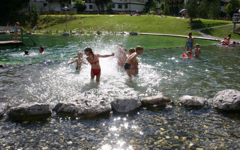 Kinder spielen und planschen in einem flachen Teich in der Nähe eines grasbewachsenen Hügels, mit Gebäuden im Hintergrund an einem sonnigen Tag.