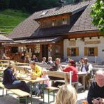 An einem sonnigen Tag sitzen Menschen an Holztischen im Freien und essen und trinken vor einem rustikalen Holzgebäude.