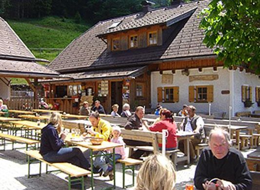 An einem sonnigen Tag sitzen Menschen an Holztischen im Freien und essen und trinken vor einem rustikalen Holzgebäude.