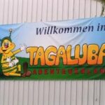 Auf dem Banner steht „Willkommen im TAGALUBA ABENTEUERLAND“ und eine Zeichentrickfigur, eine Biene, sitzt winkend im Gras und ist an einer Wellblechwand befestigt.
