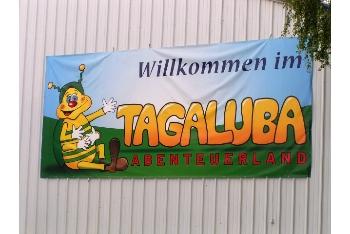 Auf dem Banner steht „Willkommen im TAGALUBA ABENTEUERLAND“ und eine Zeichentrickfigur, eine Biene, sitzt winkend im Gras und ist an einer Wellblechwand befestigt.