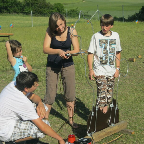 Vier Kinder spielen im Freien ein selbstgebautes Drahtschleifenspiel in Form einer Person, mit Gras, Bäumen und einem Zaun im Hintergrund.