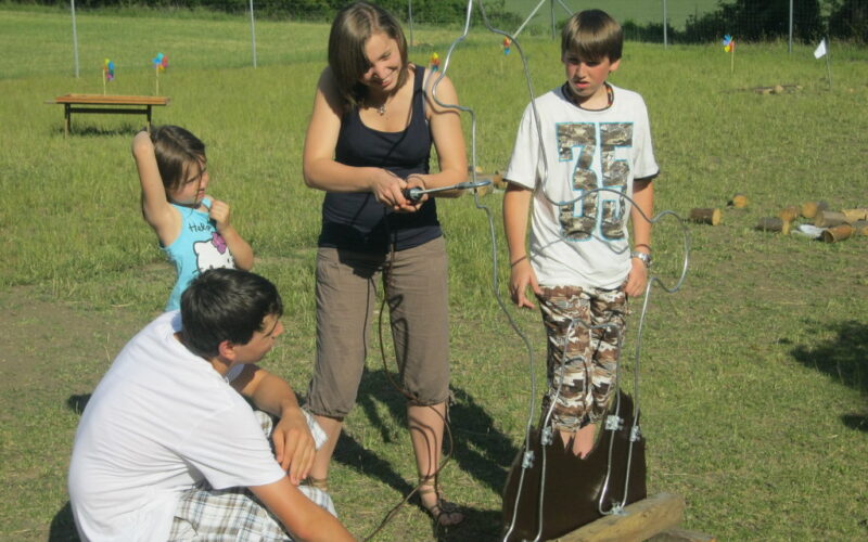 Vier Kinder spielen im Freien ein selbstgebautes Drahtschleifenspiel in Form einer Person, mit Gras, Bäumen und einem Zaun im Hintergrund.