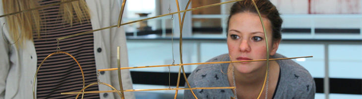 Eine Frau untersucht aufmerksam eine geometrische Metallskulptur, während im Hintergrund eine weitere Person in der Nähe steht.