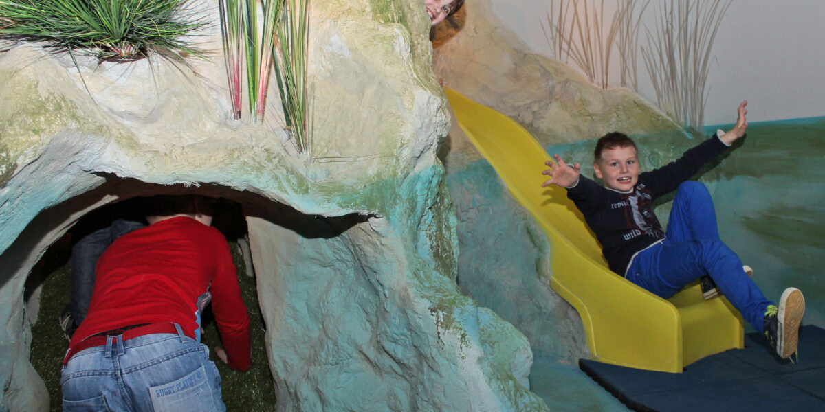 Drei Kinder spielen auf einem Indoor-Spielplatz mit einem Tunnel und einer gelben Rutsche; ein Kind krabbelt in den Tunnel, während zwei andere aus der Rutsche kommen.