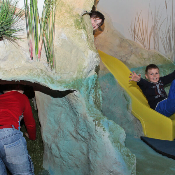 Drei Kinder spielen auf einem Indoor-Spielplatz mit einem Tunnel und einer gelben Rutsche; ein Kind krabbelt in den Tunnel, während zwei andere aus der Rutsche kommen.