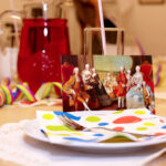 Ein gedeckter Tisch mit einer bunten Serviette, einer Gabel und einem Glas, auf dem eine Karte mit einem klassischen Gemälde von Aristokraten zu sehen ist. Im Hintergrund sind Luftschlangen und ein Krug mit rotem Getränk zu sehen.