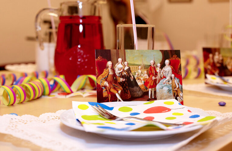 Ein gedeckter Tisch mit einer bunten Serviette, einer Gabel und einem Glas, auf dem eine Karte mit einem klassischen Gemälde von Aristokraten zu sehen ist. Im Hintergrund sind Luftschlangen und ein Krug mit rotem Getränk zu sehen.