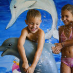 Zwei Kinder in Badeanzügen spielen und lachen mit einer Delfinstatue vor einer Wandmalerei, die Tiere des Meeres zeigt.