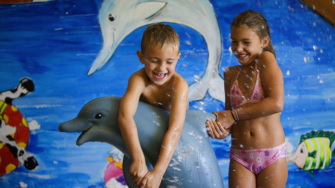 Zwei Kinder in Badeanzügen spielen und lachen mit einer Delfinstatue vor einer Wandmalerei, die Tiere des Meeres zeigt.