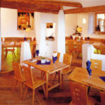 Ein modernes Restaurant mit Holztischen und -stühlen, weißen Wänden, freiliegenden Holzbalken und säuberlich gefalteten Servietten auf jedem Tisch.