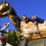 Eine Familie posiert im Freien mit zwei Kindern, die auf eine große hölzerne Pferdeskulptur klettern und darauf sitzen, vor einem klaren blauen Himmel.