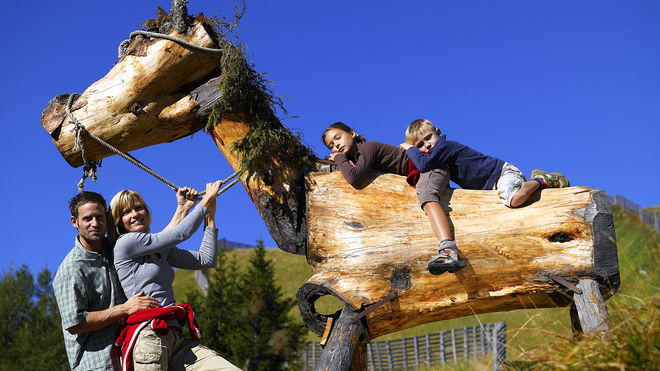 Eine Familie posiert im Freien mit zwei Kindern, die auf eine große hölzerne Pferdeskulptur klettern und darauf sitzen, vor einem klaren blauen Himmel.