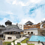 Ein moderner Ferienkomplex in den Bergen mit mehreren Gebäuden, einem Außenpool, Liegestühlen und bewaldeten Hügeln im Hintergrund unter einem teilweise bewölkten Himmel.